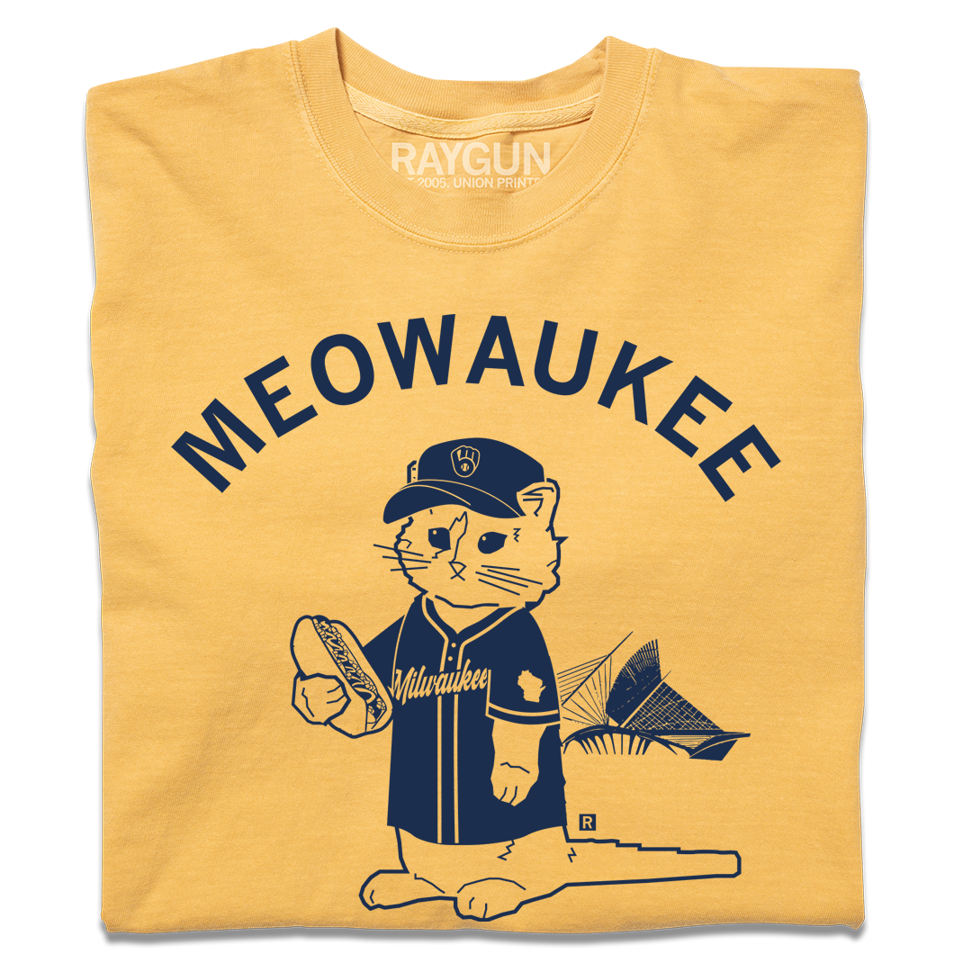 A folded mustard yellow t-shirt with navy ink that says, "MEOWAUKEE" in curved text over Gary The Cat wearing Brewers gear while holding a hotdog.