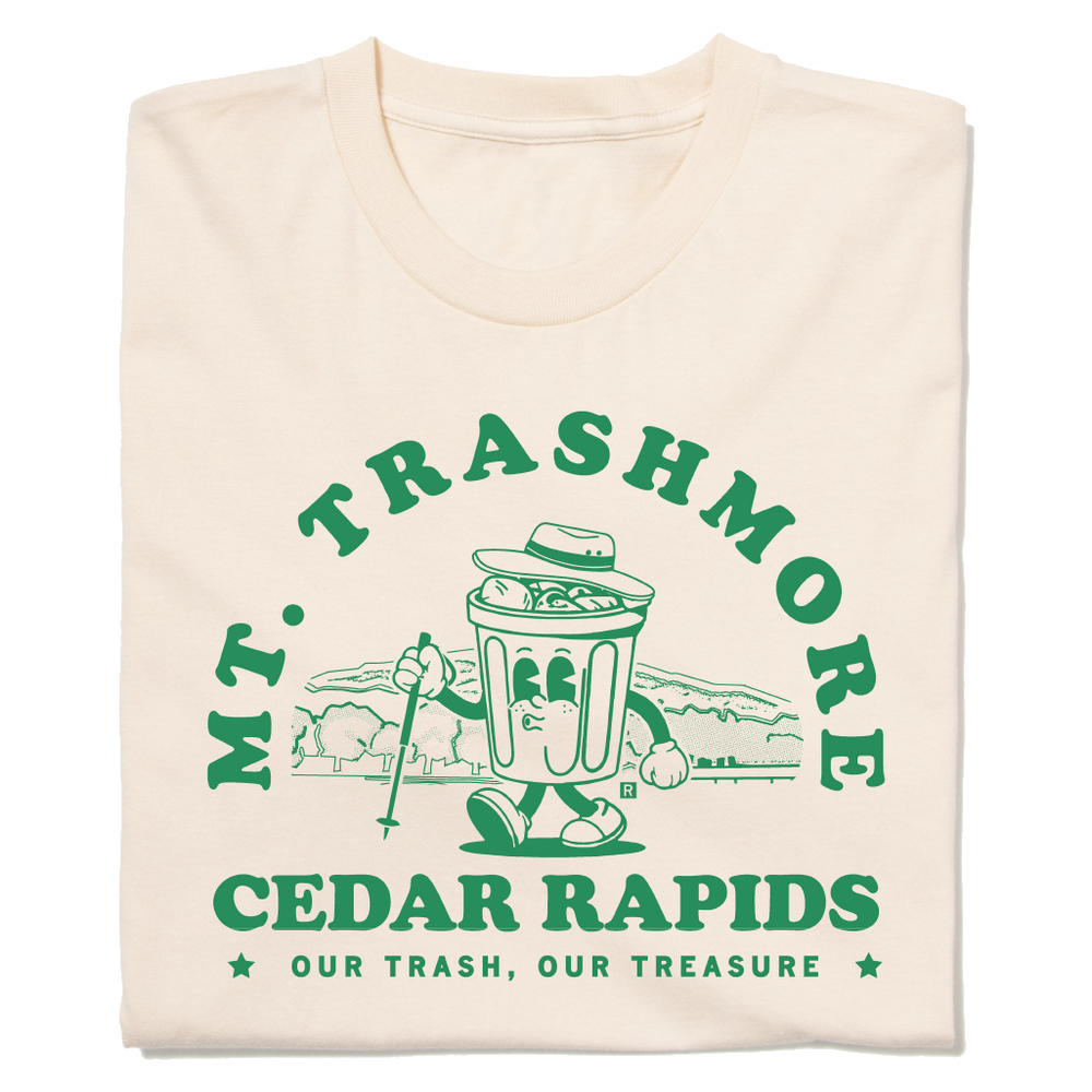 A folded cream shirt with green ink that reads "Mt. Trashmore, Cedar Rapids. Our trash, our treasure" framing an image of a cartoon trashcan going for a hike with Mt. Trashmore in the background.