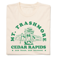 A folded cream shirt with green ink that reads "Mt. Trashmore, Cedar Rapids. Our trash, our treasure" framing an image of a cartoon trashcan going for a hike with Mt. Trashmore in the background.
