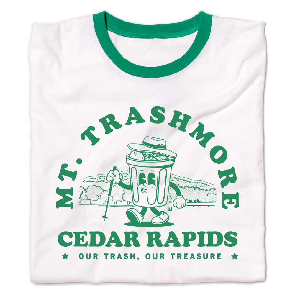 A folded white ringer shirt with green accents and green ink that reads "Mt. Trashmore, Cedar Rapids. Our trash, our treasure" framing an image of a cartoon trashcan going for a hike with Mt. Trashmore in the background.
