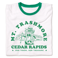 A folded white ringer shirt with green accents and green ink that reads "Mt. Trashmore, Cedar Rapids. Our trash, our treasure" framing an image of a cartoon trashcan going for a hike with Mt. Trashmore in the background.