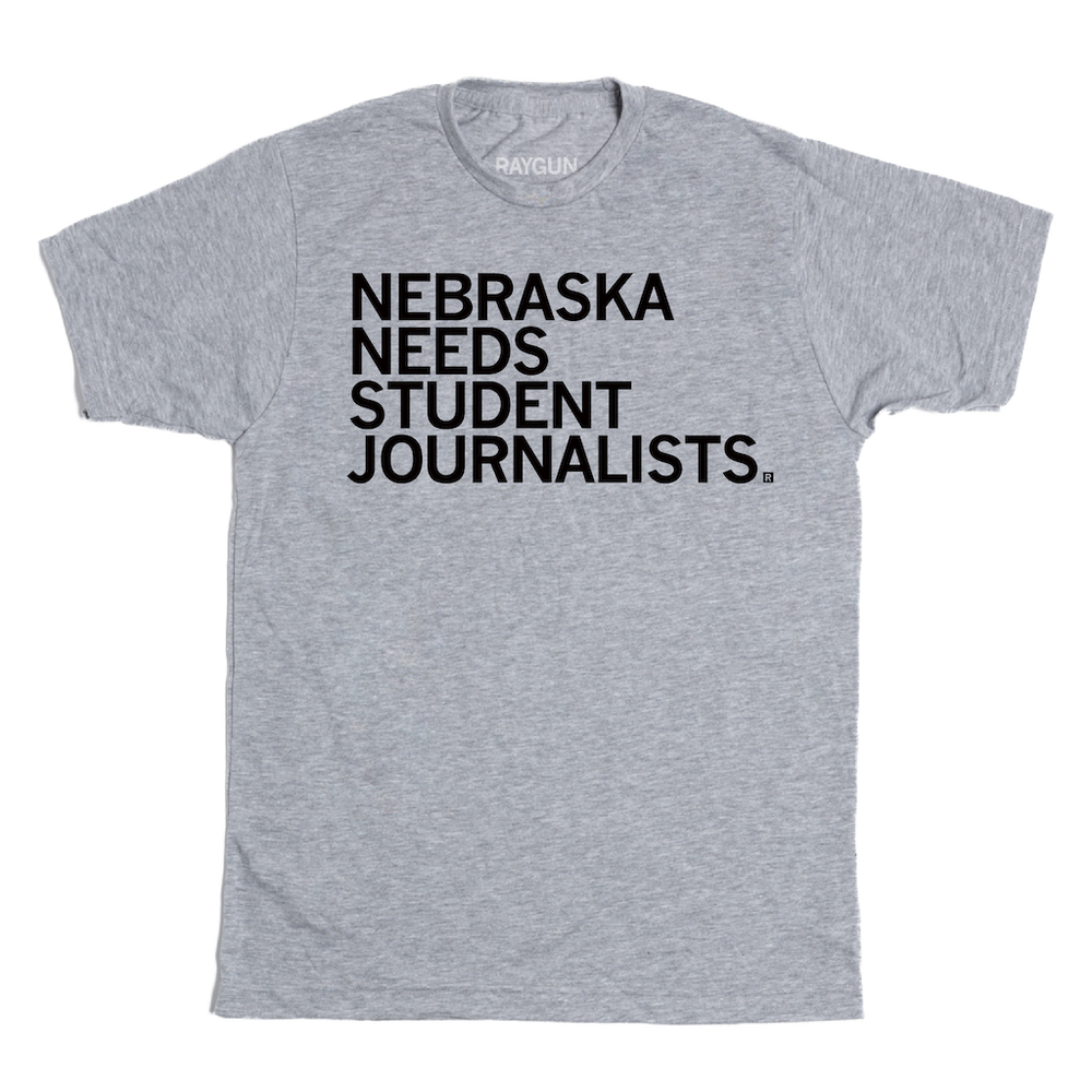 A heather grey short sleeved t-shirt with left aligned black in text that reads "Nebraska Needs Student Journalists"