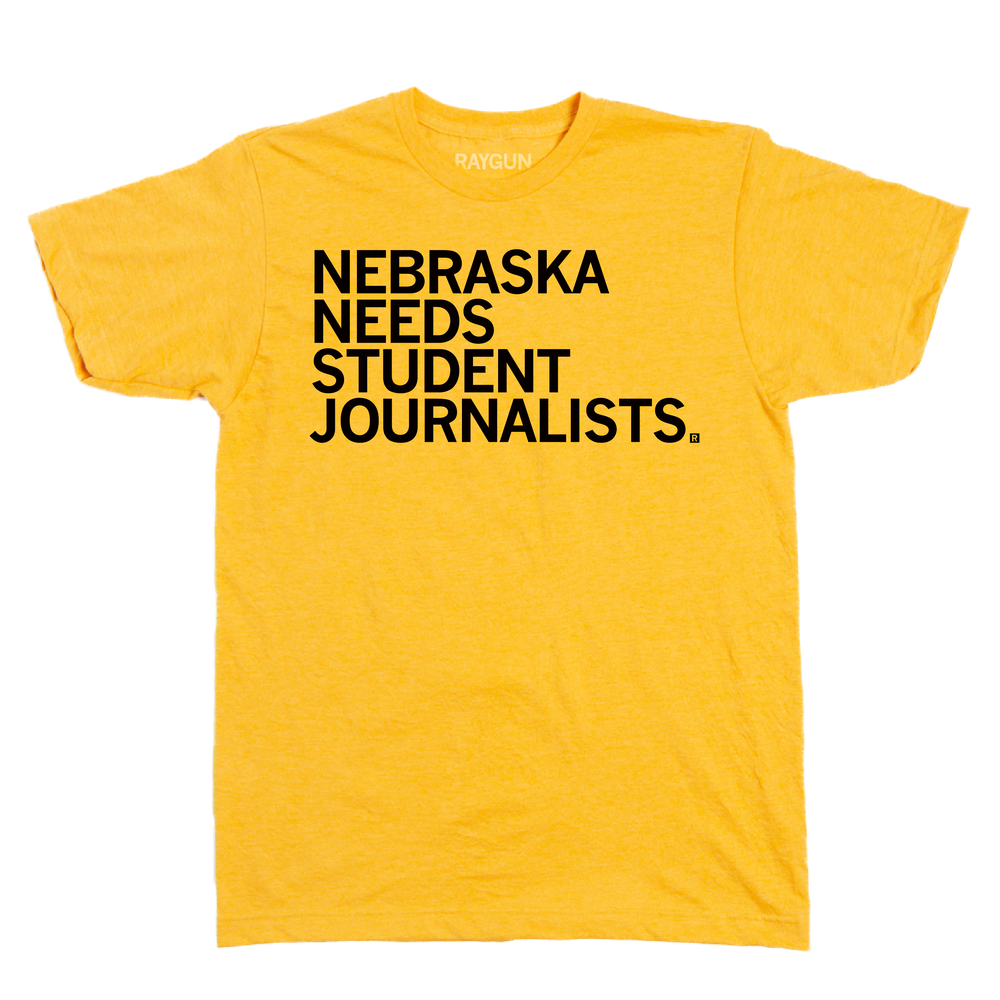 A heather mellow yellow short sleeved t-shirt with left aligned black in text that reads "Nebraska Needs Student Journalists"