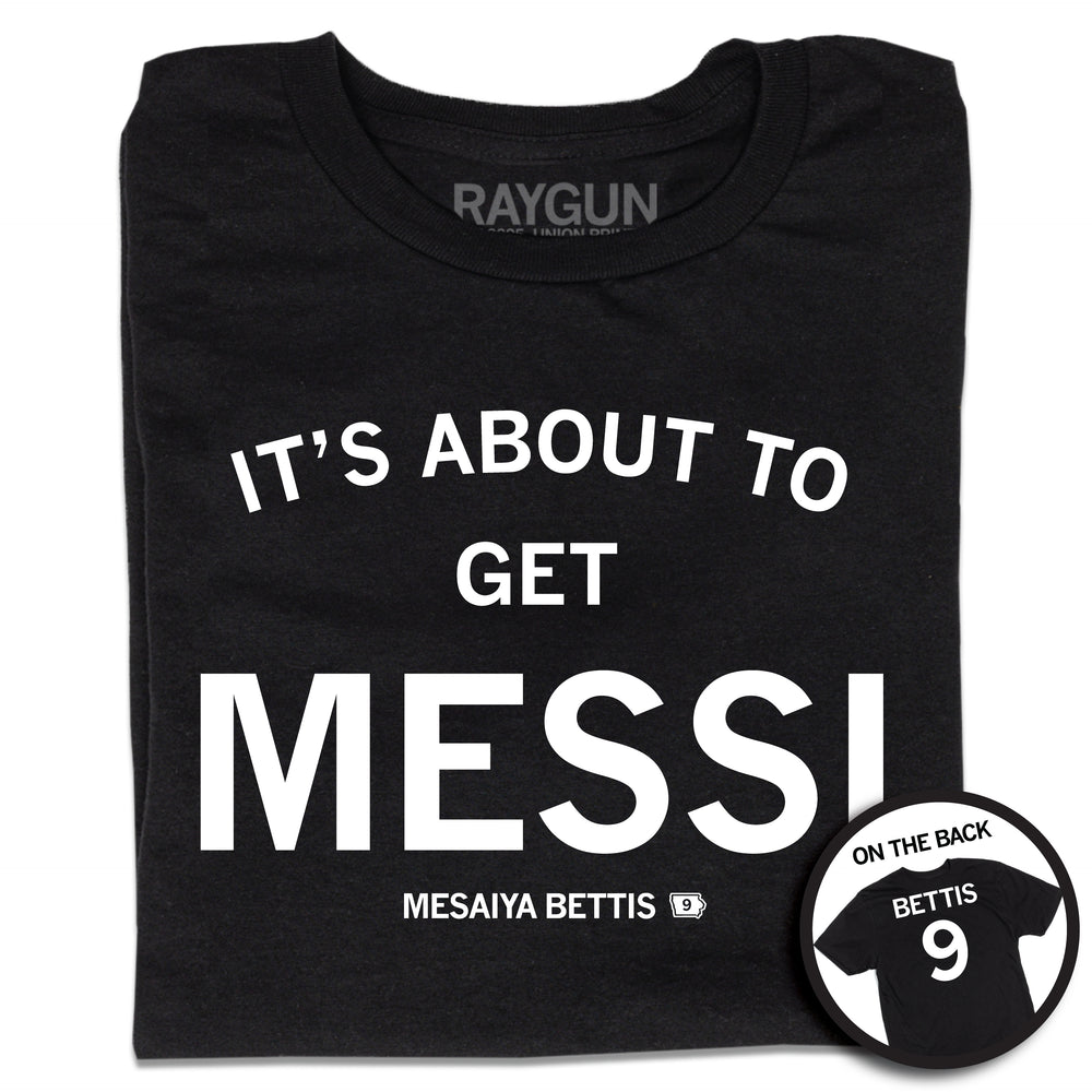 A folded short sleeve black t-shirt with centered white ink print that reads "It's About to Get Messi" with smaller text below that says, "Mesaiya Bettis" beside a small outline of Iowa with the number 9 in it. A second picture in a small circle frame is in the bottom right corner, showing the back print. The back is laid out like a jersey with "Bettis" printed across the shoulders above the number 9.