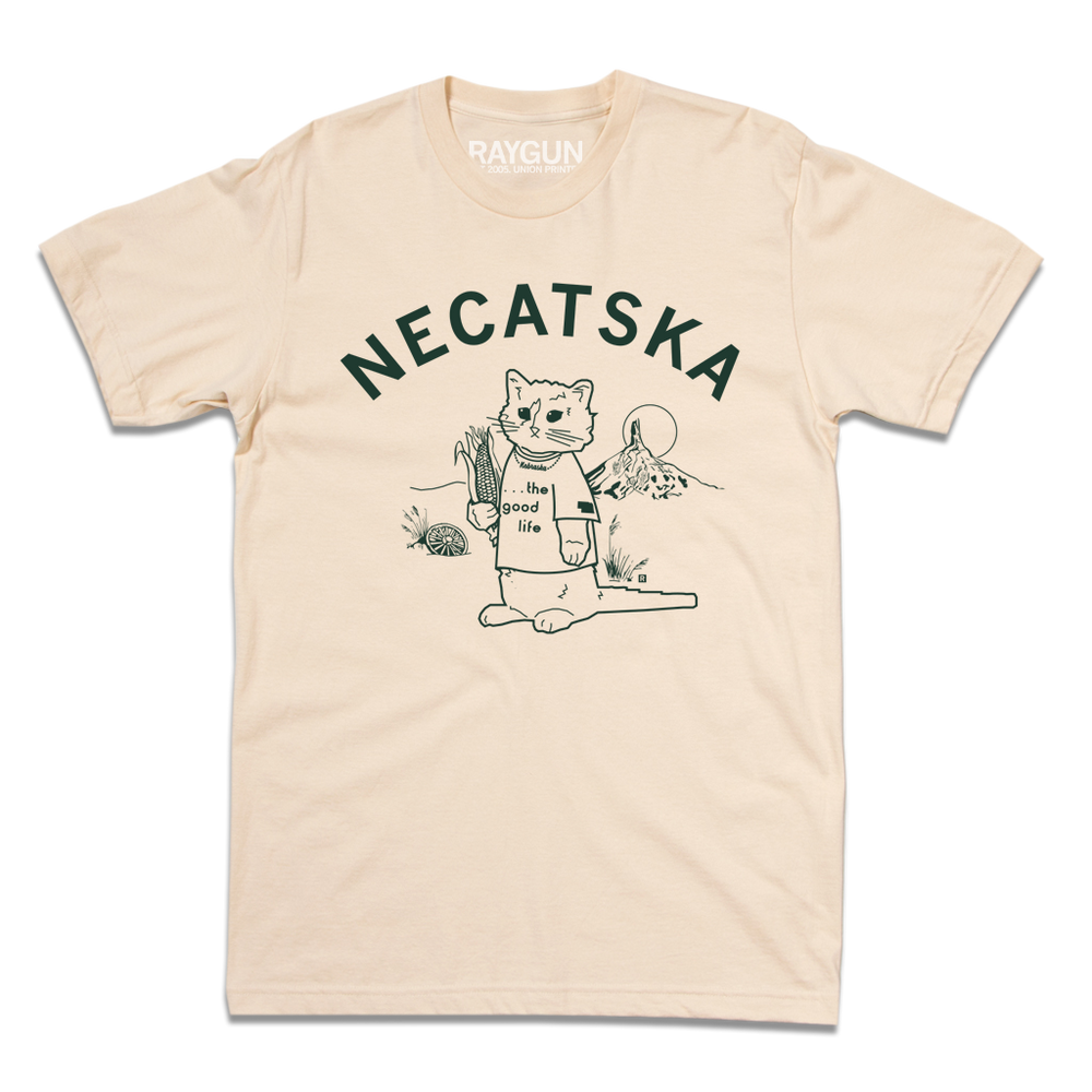 An off white shirt with dark green ink. Curved text across the top says, "Necatska" above an illustration of RAYGUN's cat mascot wearing a t-shirt that reads "the good life" along with a Nebraska necklace. He's holding on ear of corn in his right end and rural Nebraska landscape is behind him.