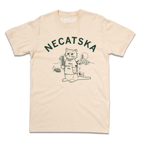 An off white shirt with dark green ink. Curved text across the top says, "Necatska" above an illustration of RAYGUN's cat mascot wearing a t-shirt that reads "the good life" along with a Nebraska necklace. He's holding on ear of corn in his right end and rural Nebraska landscape is behind him.