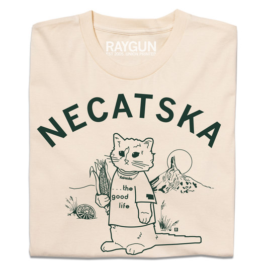 A folded cream t-shirt with dark green ink. Curved text across the top says, "Necatska" above an illustration of RAYGUN's cat mascot wearing a t-shirt that reads "the good life" along with a Nebraska necklace. He's holding on ear of corn in his right end and rural Nebraska landscape is behind him.