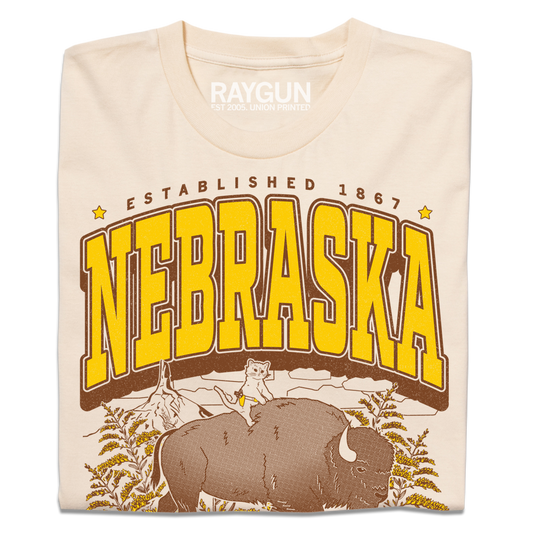 Cream-colored Nebraska t-shirt with a graphic of a cat riding a bison, featuring the text ESTABLISHED 1867 and NEBRASKA in large yellow and brown letters.