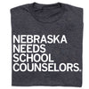 Charcoal gray RAYGUN t-shirt with white text that reads NEBRASKA NEEDS SCHOOL COUNSELORS.