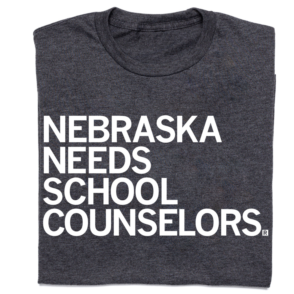 Charcoal gray RAYGUN t-shirt with white text that reads NEBRASKA NEEDS SCHOOL COUNSELORS.