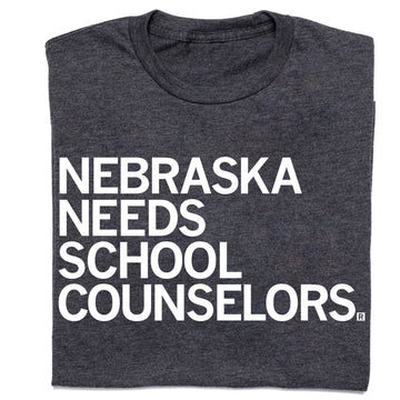 Charcoal gray RAYGUN t-shirt with white text that reads NEBRASKA NEEDS SCHOOL COUNSELORS.