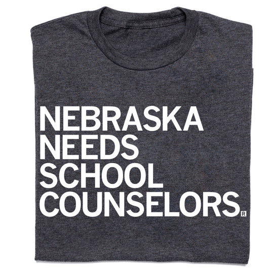 Charcoal gray RAYGUN t-shirt with white text that reads NEBRASKA NEEDS SCHOOL COUNSELORS.