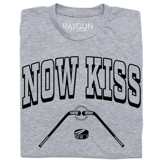 A folded heather grey shirt with blank ink. Curved text reads "Now Kiss" above an illustration of two hocky sticks  touching with a puck beneath.