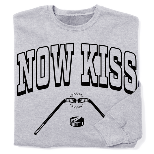 A folded light grey sweatshirt with blank ink print that reads "Now Kiss" above an illustration of two hockey sticks touching above a puck.