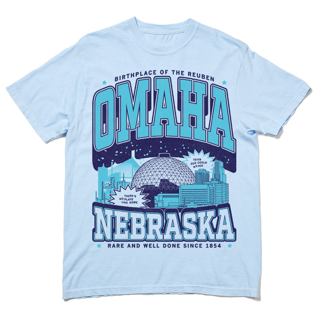 Light blue garment-dyed heavyweight cotton T-shirt featuring an Omaha, Nebraska skyline graphic with the text Birthplace of the Reuben, Your zoo could never, Theres no place like dome, and Rare and well done since 1854.