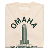 Cream-colored Omaha t-shirt featuring a dark green graphic of a skyscraper, the word OMAHA arched above it, and the text WE KNOW WHAT IT printed below.