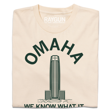 Cream-colored Omaha t-shirt featuring a dark green graphic of a skyscraper, the word OMAHA arched above it, and the text WE KNOW WHAT IT printed below.