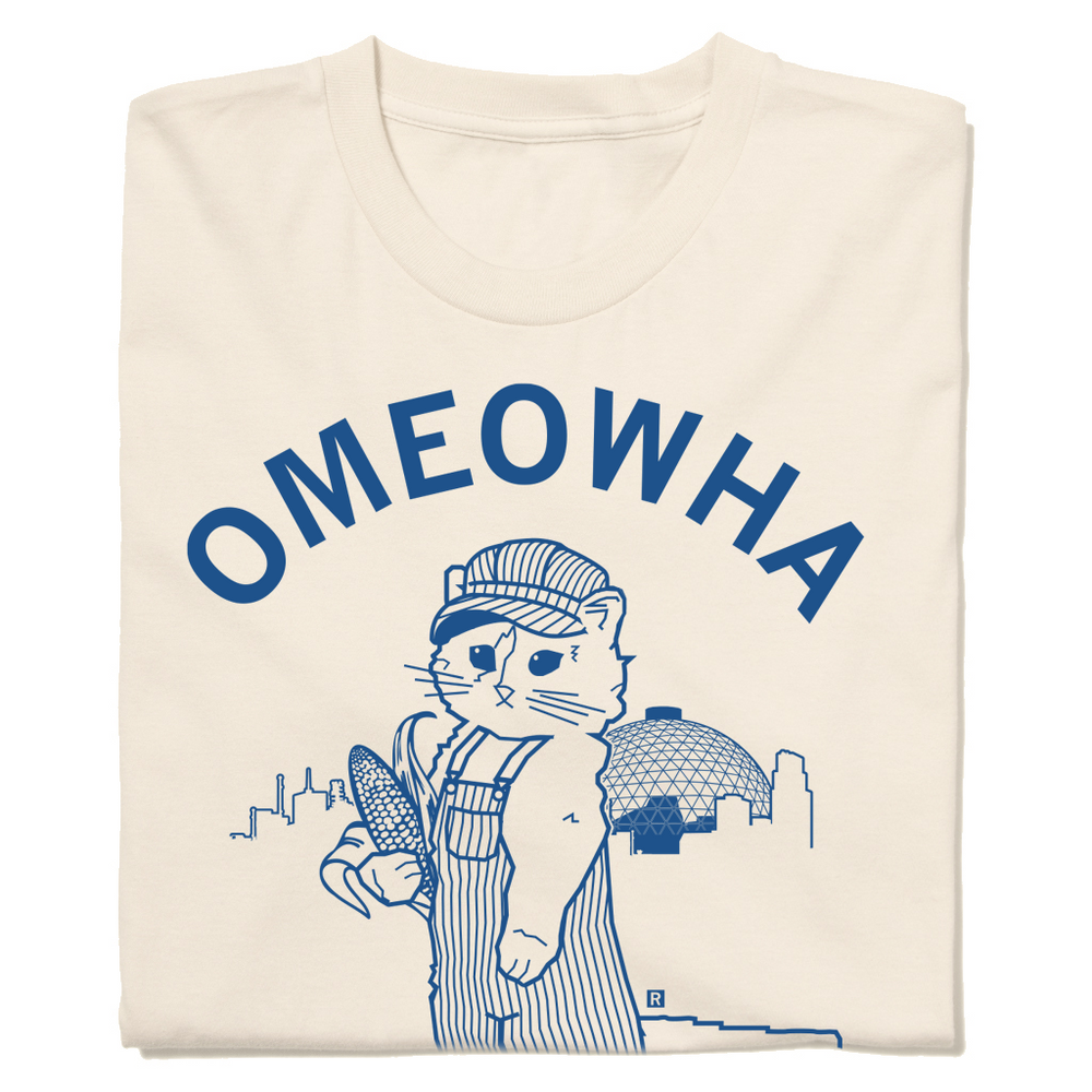 Folded natural shirt with royal ink that says, "Omeowha" over an illustration of the Omaha skyline and dome of the Omaha zoo behind a cat dressed as a train conductor holding an  ear of corn.