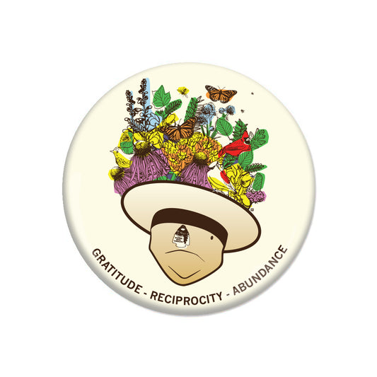A round gratitude button featuring a cartoon character wearing a hat overflowing with flowers, butterflies, and birds, with the text GRATITUDE - RECIPROCITY - ABUNDANCE curved along the bottom.