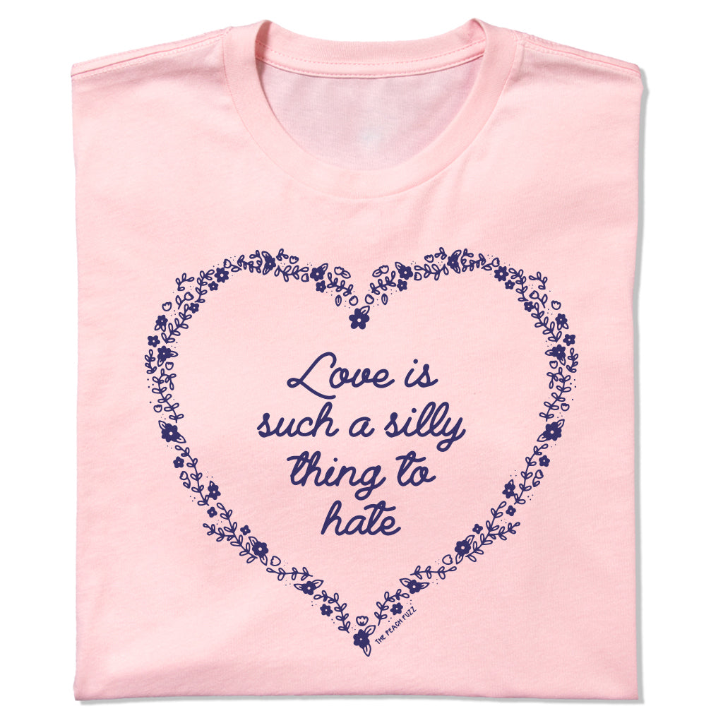 A light pink RAYGUN t-shirt featuring a dark blue floral heart outline surrounding the text Love is such a silly thing to hate.