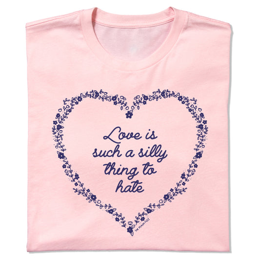 A light pink RAYGUN t-shirt featuring a dark blue floral heart outline surrounding the text Love is such a silly thing to hate.