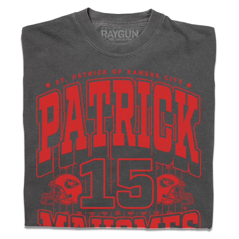 A folded asphalt shirt with bright red ink that reads "Saint Patrick of Kansas City. Patrick Mahomes #15. There's No Place Like Mahomes" over an illustration of a football field with a football helmet to either side.