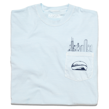 A folded light blue pocket t-shirt with navy ink. An illustration of the Chicago skyline is printed above the pocket with the Cloud Gate statue printed on the pocket.
