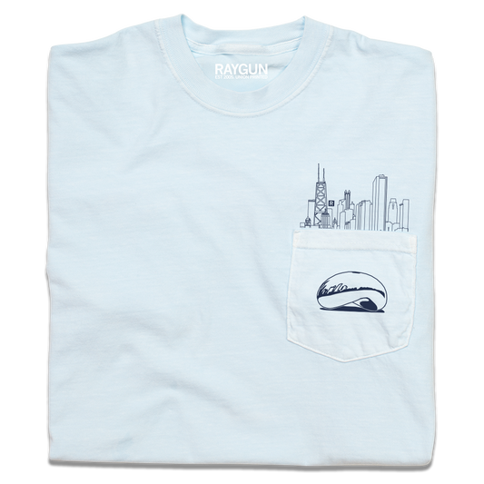 A folded light blue pocket t-shirt with navy ink. An illustration of the Chicago skyline is printed above the pocket with the Cloud Gate statue printed on the pocket.