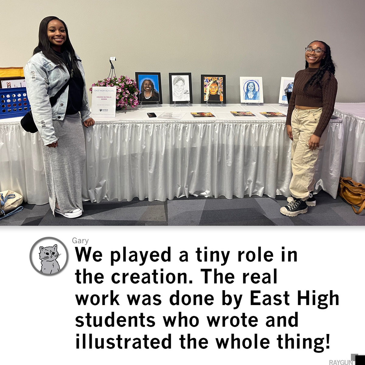 Two young women stand on either side of a table displaying framed portrait artwork of Waterloo Women at an East High School event.