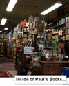 A man in a heavyweight ivory t-shirt and a baseball cap sits at a cluttered wooden desk in Paul’s Book Store, surrounded by tall, packed bookshelves and a rolling library ladder.