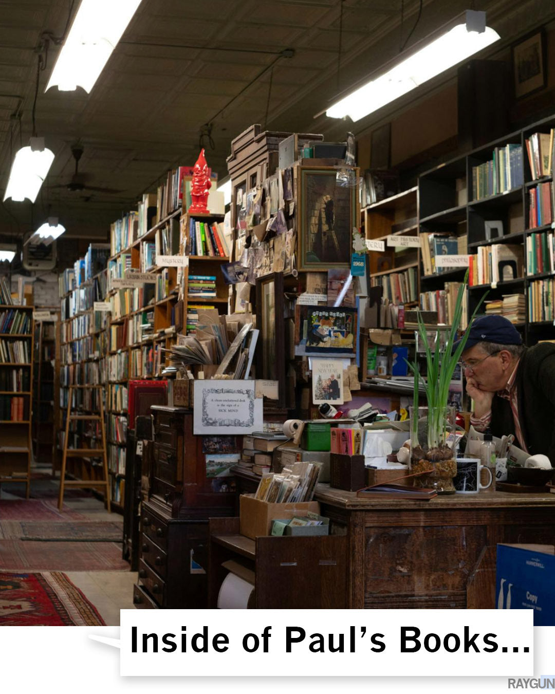 A man wearing a Madison t-shirt sits at a cluttered desk in a bookstore filled with floor-to-ceiling shelves packed with books.
