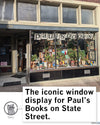 The storefront of Pauls Books on State Street, with books and plants arranged in the display window below a banner reading pauls books, showing a Madison t-shirt and Wisconsin Public Radio gear.