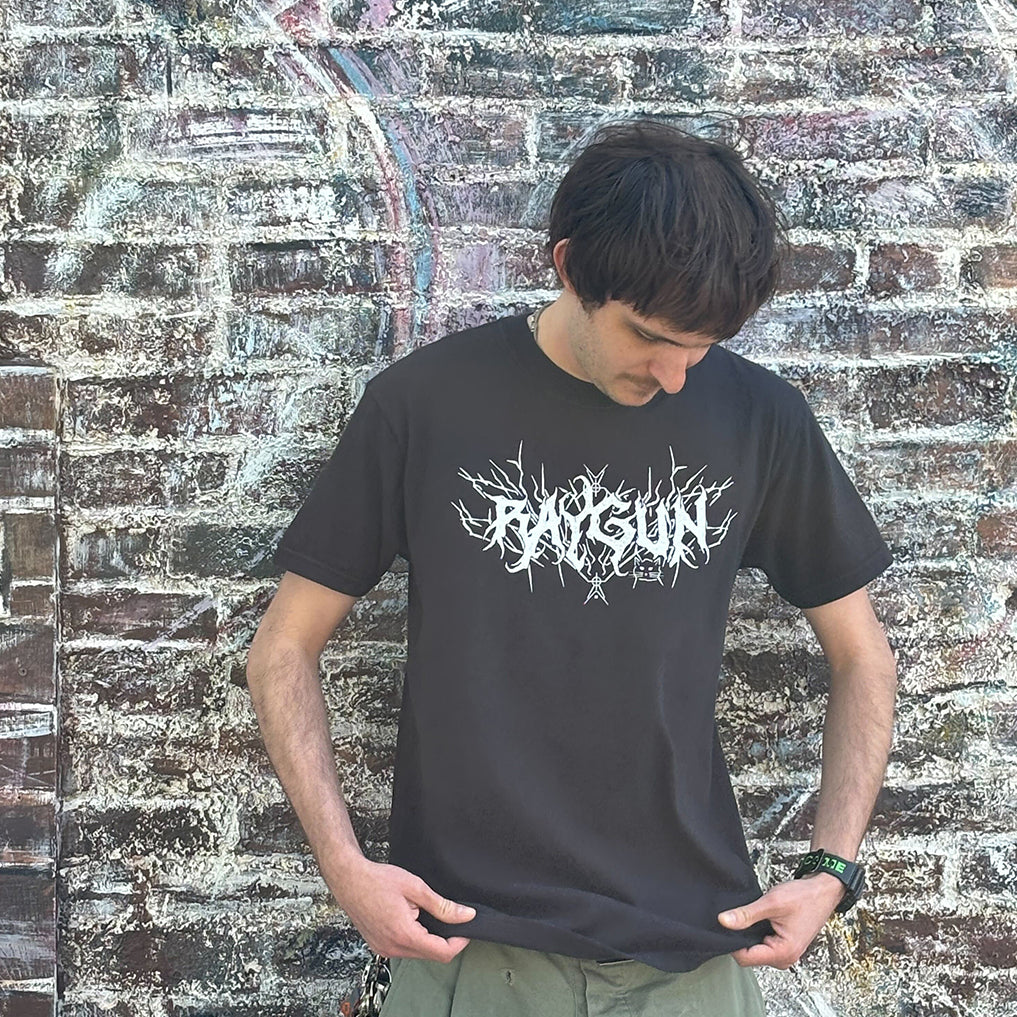 A person wearing a heavyweight black RAYGUN t-shirt with the brand name in stylized, spiky white lettering.