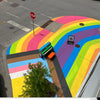 An upright piano painted with colorful designs sits on a Des Moines street corner adorned with a large Pride flag mural.