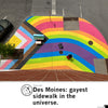 A person in a red dress and a pride T-shirt walks across a vibrant rainbow-colored crosswalk in Des Moines, passing a decorated upright piano and a black handbag sitting on the pavement.