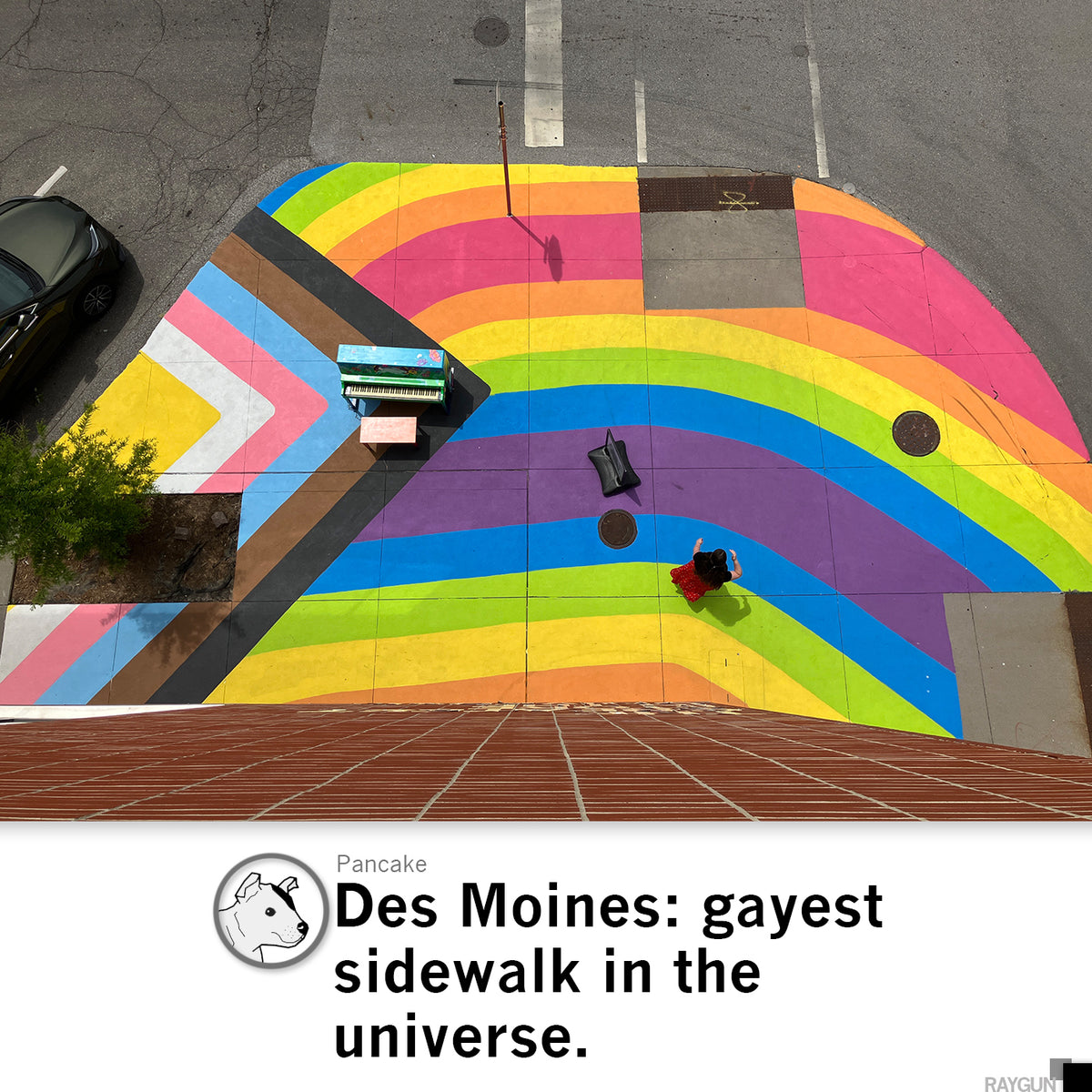 A person in a red dress and a pride T-shirt walks across a vibrant rainbow-colored crosswalk in Des Moines, passing a decorated upright piano and a black handbag sitting on the pavement.