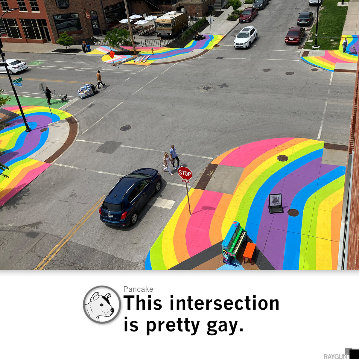 An aerial view of a Des Moines city intersection with rainbow-painted crosswalks and a colorful piano, where a person in a Pride T-shirt stands on the corner.
