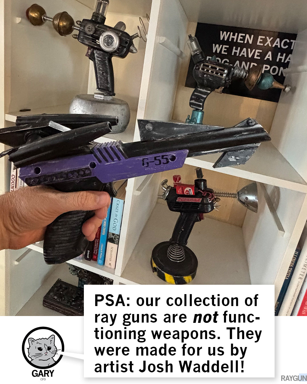 A hand holds a purple and black G-550 prop ray gun in front of a white shelf displaying several other handmade ray gun models, with a text overlay from Gary the cat stating these are non-functioning art pieces by Josh Waddell.
