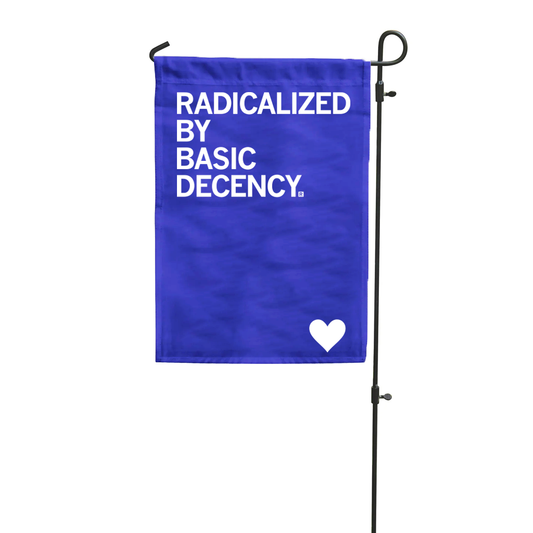 A blue RAYGUN garden flag on a metal stand displays the white text RADICALIZED BY BASIC DECENCY with a small white heart at the bottom.