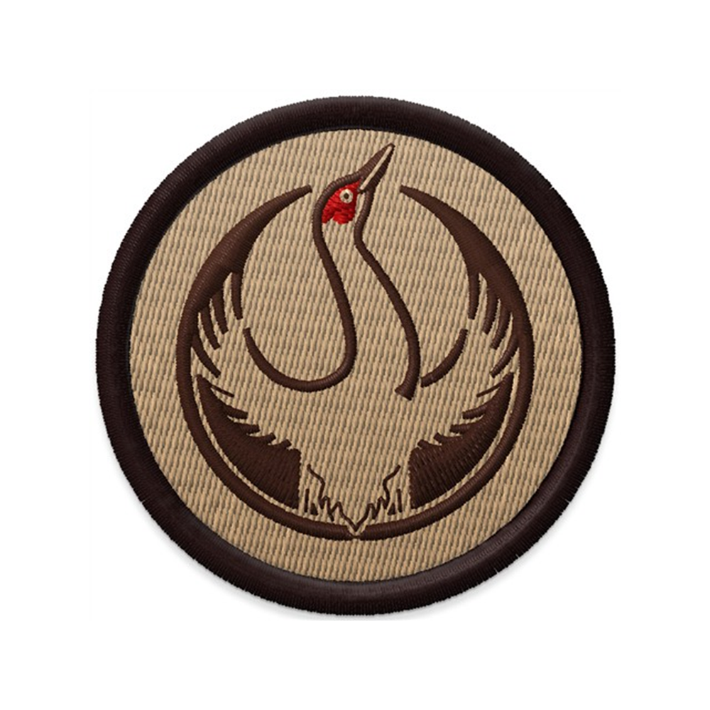 An embroidered circular patch with the Rebel Sandhill Crane Alliance Logo. It uses a rebel sandhill crane with outstretched wings to mimic the shape of the Star Wars Rebel Alliance Logo. The bird is printed in red and brown. The edge of the patch i s dark brown and the background is tan.