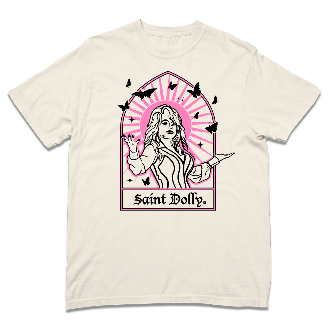 Comfort Colors ivory heavyweight t-shirt with pink & black print depicting Dolly Parton with her arms outstretched. A stained glass frame is around her, with a pink sun behind her, black butterflies around her, and text at the bottom that says, "SAINT DOLLY."