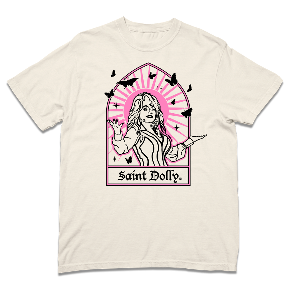 Comfort Colors ivory heavyweight t-shirt with pink & black print depicting Dolly Parton with her arms outstretched. A stained glass frame is around her, with a pink sun behind her, black butterflies around her, and text at the bottom that says, "SAINT DOLLY."