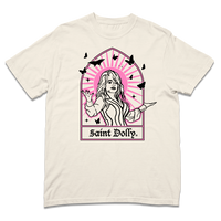 Comfort Colors ivory heavyweight t-shirt with pink & black print depicting Dolly Parton with her arms outstretched. A stained glass frame is around her, with a pink sun behind her, black butterflies around her, and text at the bottom that says, "SAINT DOLLY."