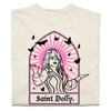 A folded off-white heavyweight t-shirt with pink & black print depicting Dolly Parton with her arms outstretched. A stained glass frame is around her, with a pink sun behind her, black butterflies around her, and text at the bottom that says, "SAINT DOLLY."