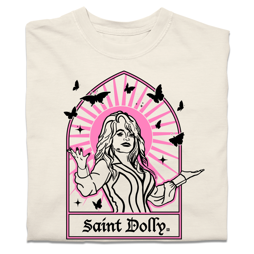 A folded off-white heavyweight t-shirt with pink & black print depicting Dolly Parton with her arms outstretched. A stained glass frame is around her, with a pink sun behind her, black butterflies around her, and text at the bottom that says, "SAINT DOLLY."
