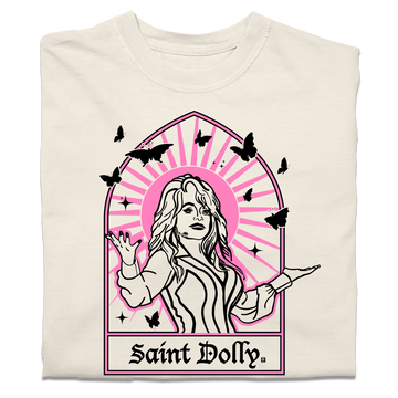 A folded off-white heavyweight t-shirt with pink & black print depicting Dolly Parton with her arms outstretched. A stained glass frame is around her, with a pink sun behind her, black butterflies around her, and text at the bottom that says, "SAINT DOLLY."
