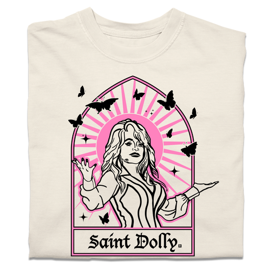 A folded off-white heavyweight t-shirt with pink & black print depicting Dolly Parton with her arms outstretched. A stained glass frame is around her, with a pink sun behind her, black butterflies around her, and text at the bottom that says, "SAINT DOLLY."