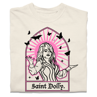 A folded off-white heavyweight t-shirt with pink & black print depicting Dolly Parton with her arms outstretched. A stained glass frame is around her, with a pink sun behind her, black butterflies around her, and text at the bottom that says, "SAINT DOLLY."