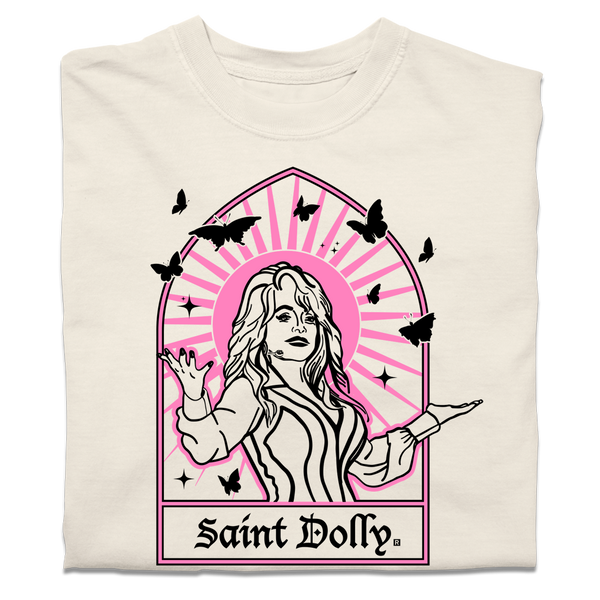 A folded off-white heavyweight t-shirt with pink & black print depicting Dolly Parton with her arms outstretched. A stained glass frame is around her, with a pink sun behind her, black butterflies around her, and text at the bottom that says, 