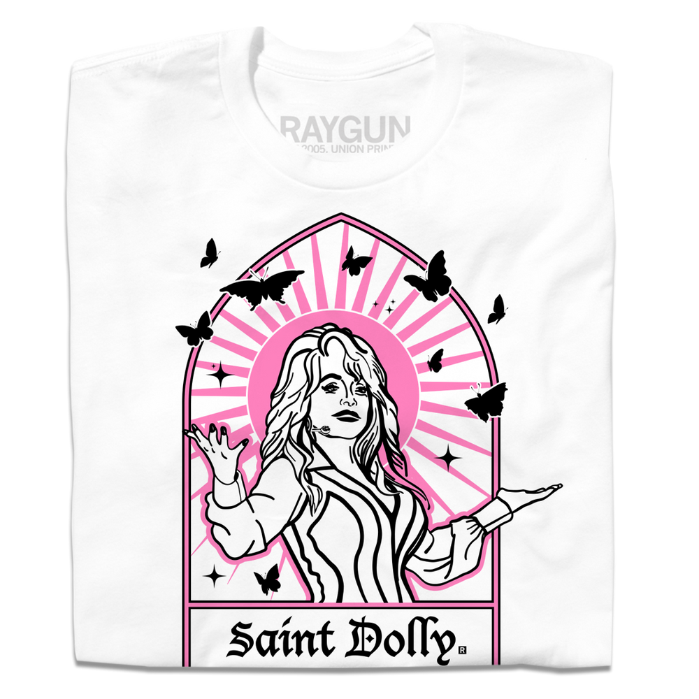 A folded white t-shirt with pink & black print depicting Dolly Parton with her arms outstretched. A stained glass frame is around her, with a pink sun behind her, black butterflies around her, and text at the bottom that says, "SAINT DOLLY."